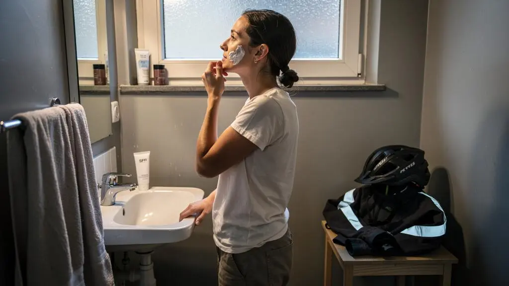 A woman applying moisturizer in a small bathroom in the morning, with her bike helmet and commuter jacket on a stool beside the sink.