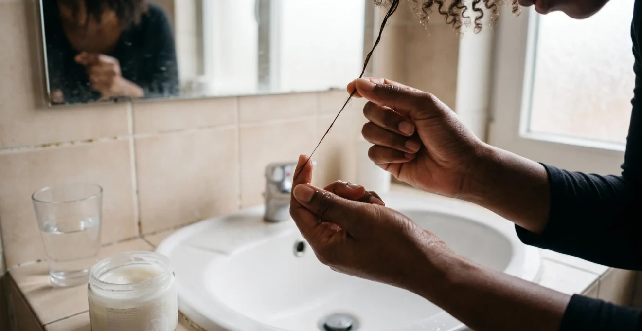 A woman stretches a wet strand of curly hair between her fingers over a bathroom sink to test its elasticity.