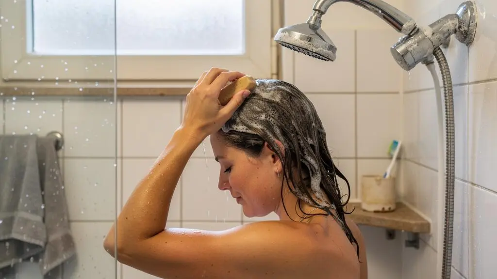 A woman washing her hair with a solid shampoo bar in a bathroom with limescale marks on the faucet and tiles.