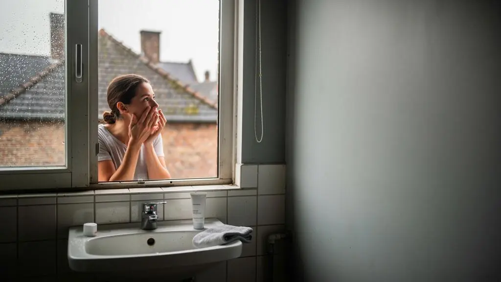 A woman applies moisturiser to her damp face at a bathroom sink in soft morning light, with an unbranded skincare tube on the sink edge.