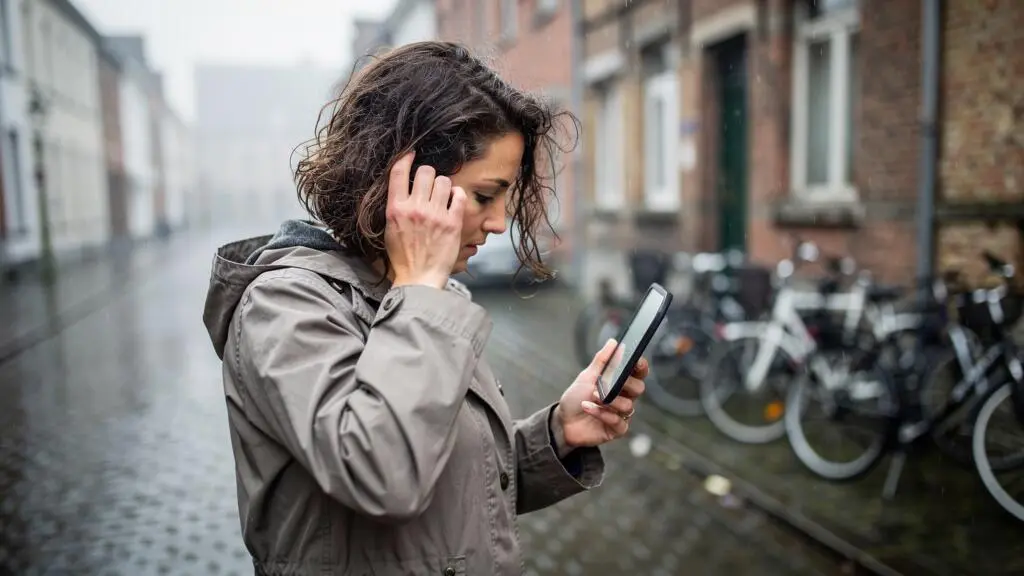 A woman on a damp Belgian street checks frizzed, flyaway hair in a small hand mirror on a humid drizzly morning.