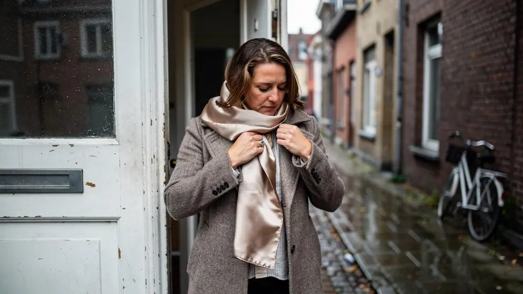 A woman wraps a satin-lined scarf around her hair at the door of her apartment before going out into a drizzly Belgian street.